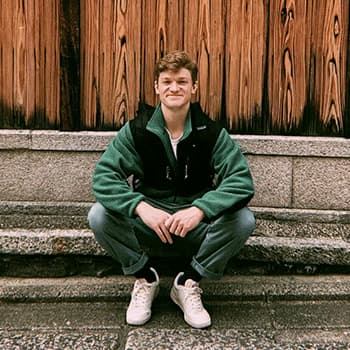 Charlie is sitting on a step. He is wearing grey jeans and a green jumper and he is smiling at the camera.