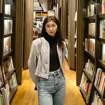 Sixian standing in between some book shelves. She has her hands behind her back and is wearing a black jumper, a grey blazer and jeans.