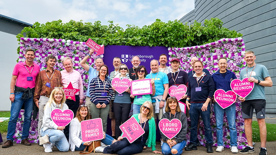 A large group of people stand and perch together in front of flowery and purple backdrops. Many of them hold up pink signs that say things such as Alumni Reunion and LboroFamily
