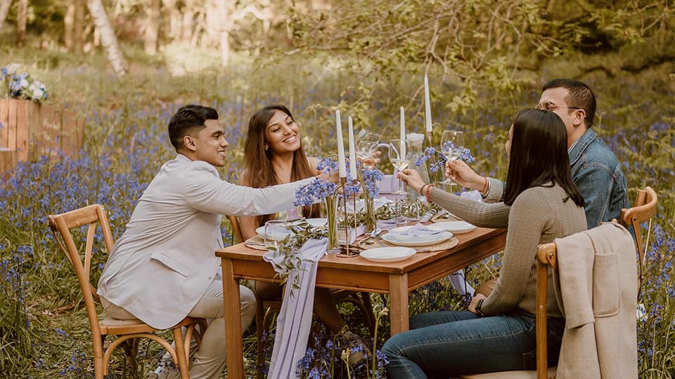 Aditya Mukherjee and Aarushi Mittal sitting at a wooden table with their friends surrounded by bluebells at Burleigh Wood, Loughborough University. They are smiling and raising their glasses.