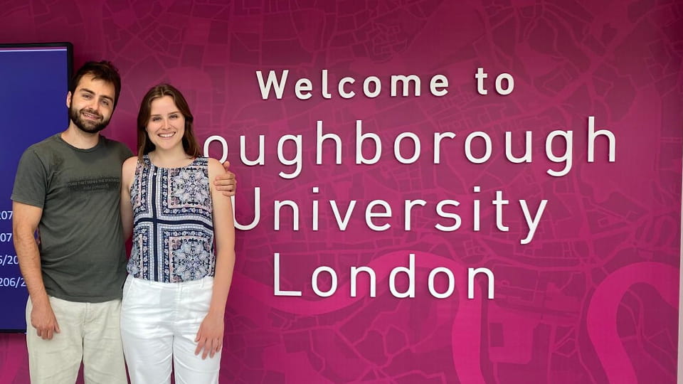 Alexandre Martins stands with someone at the entrance to the Loughborough University London campus. A welcome sign is on the pink wall behind them.
