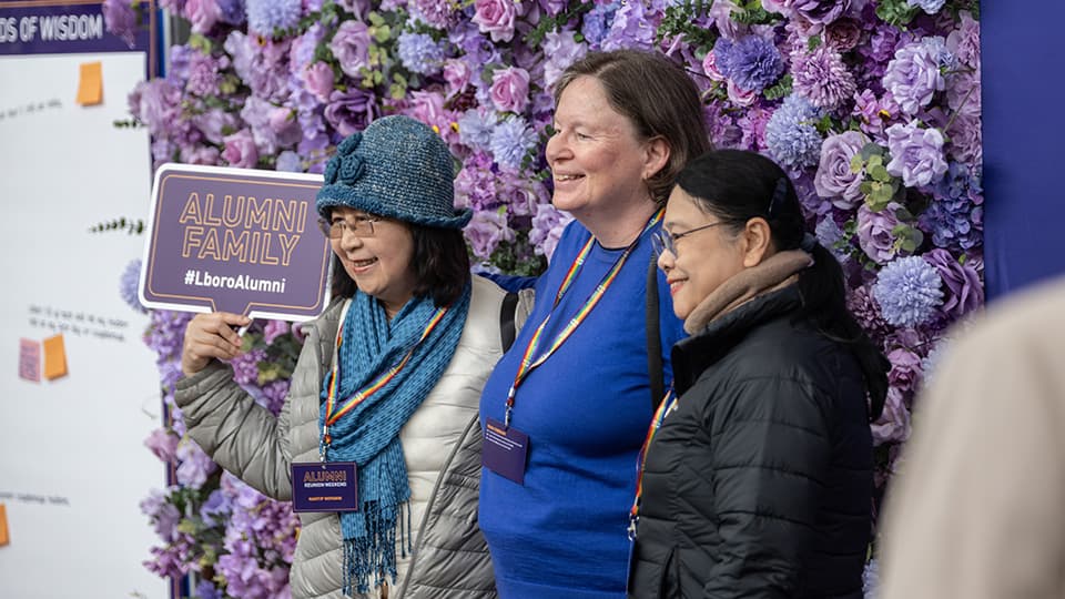 A side angle shot of three people having their photo taken. The three people are stood smiling in front of a wall of flowers. One person holds a sign that says Alumni Family.
