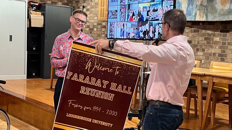 Chris Mallaband smiling as he is presented a board by Dr Ash Casey who is holding a microphone. They stand in front of a stage area.