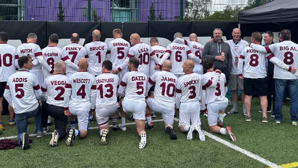 A group of football alumni wearing white football shirts with names on the back. They are standing and kneeling.