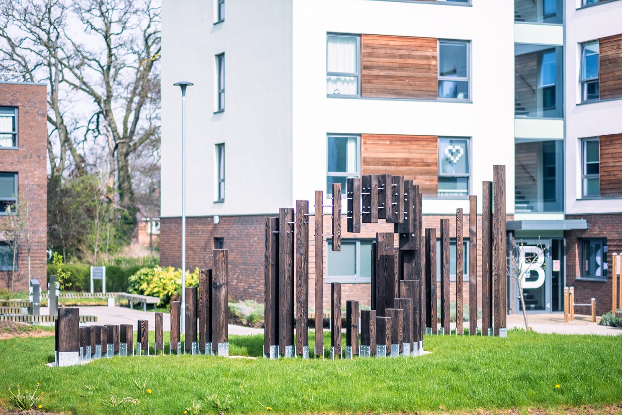 A wooden sculpture with multiple vertical panels of wood arising from a grassy mound in a wave like formation. There is a blank circular space in the middle of the sculpture.