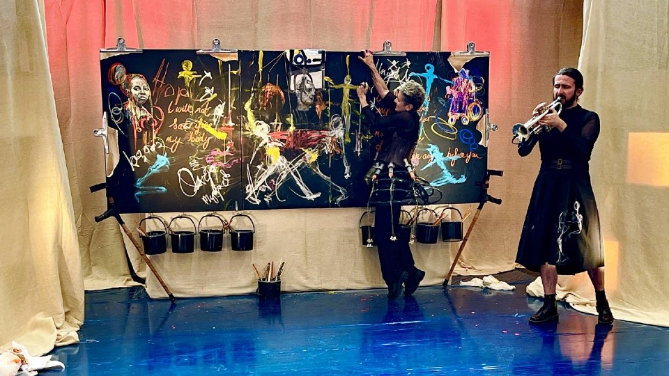 A performance scene where Rachel paints energetically on a large black canvas covered in colorful abstract figures and writing, while another person in a long black outfit plays a trumpet beside them on a glossy blue floor, with paint buckets hanging below the artwork and warm lighting behind.