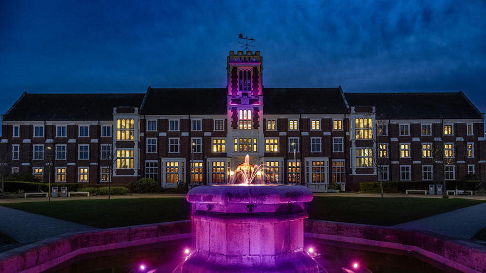 Atmospheric evening image of Hazlerigg Building behind the lawn fountain, both lit up in purple light.