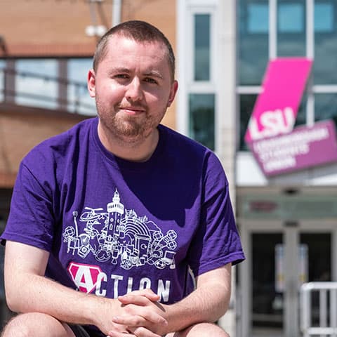 Nathan sitting outside the Loughborough Students' Union building wearing an Action t-shirt.