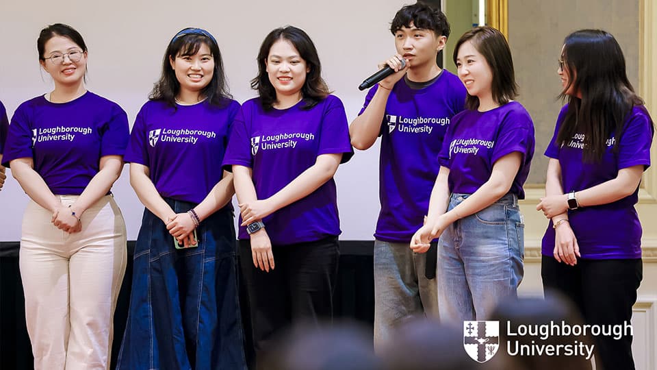Six alumni wearing purple t-shirts with the Loughborough University logo on them in Beijing. One of them is speaking into a microphone and the rest are smiling.