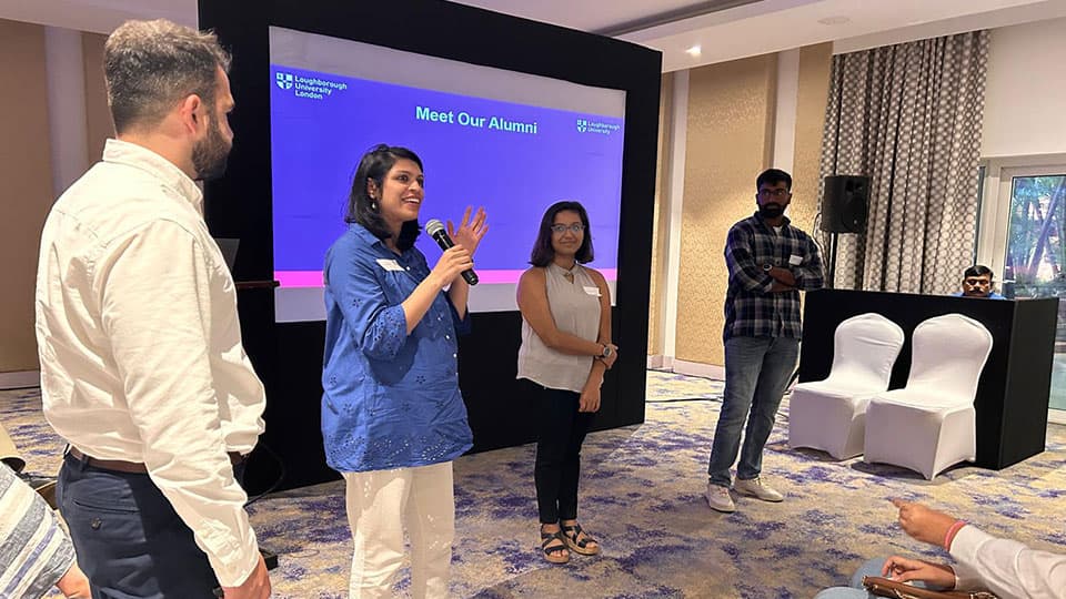 A group of three alumni standing in front of a screen that says 'Meet our alumni' talking to an audience at Loughborough University's India pre-departure event