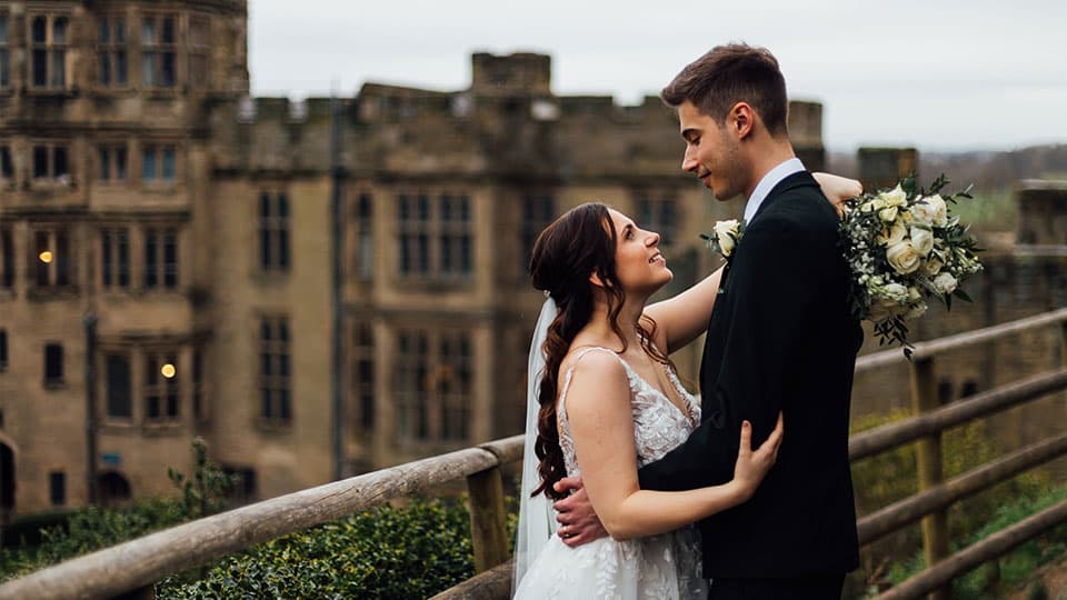 Joe and Sammy standing together looking into each others eyes on their wedding day and smiling. Warwick Castle is in the background blurred.
