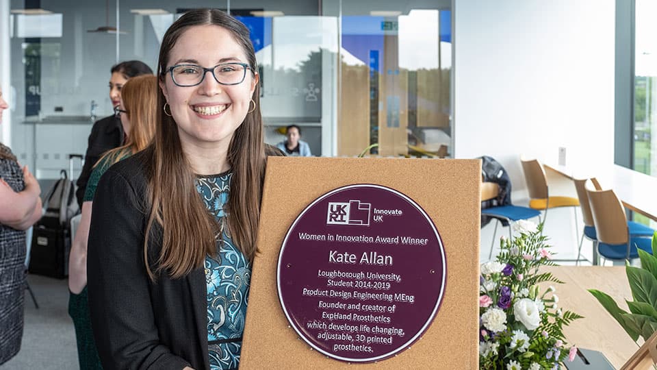Alumna Kate Allan holding a purple plaque with her name on it, she is smiling.