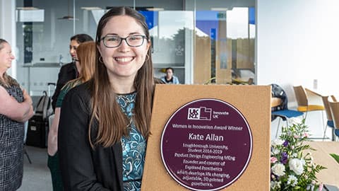 Alumna Kate Allan holding a purple plaque with her name on it, she is smiling.