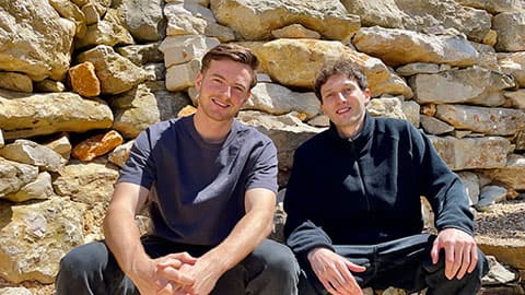 Ronan Harvey Kelly and Glenn Keller sitting next to each other smiling and they are sat on a wall with a wall behind them as the background.