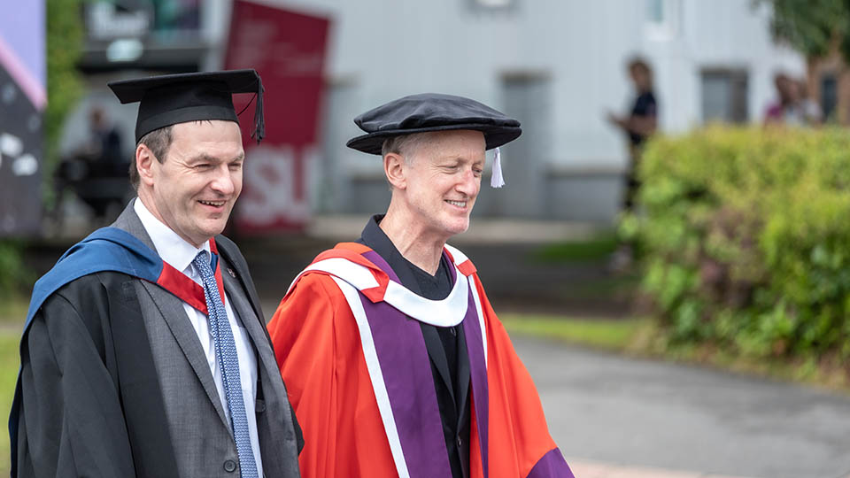Michael walking alongside a member of staff wearing graduation attire