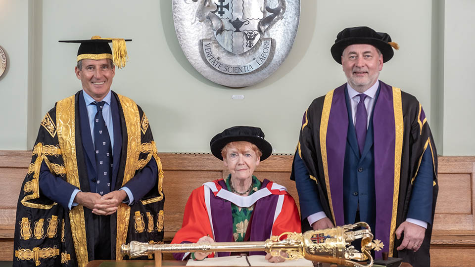 Vera sat down at a table wearing graduation attire with Lord Seb Coe and Professor Nick Jennings stood on either side of her