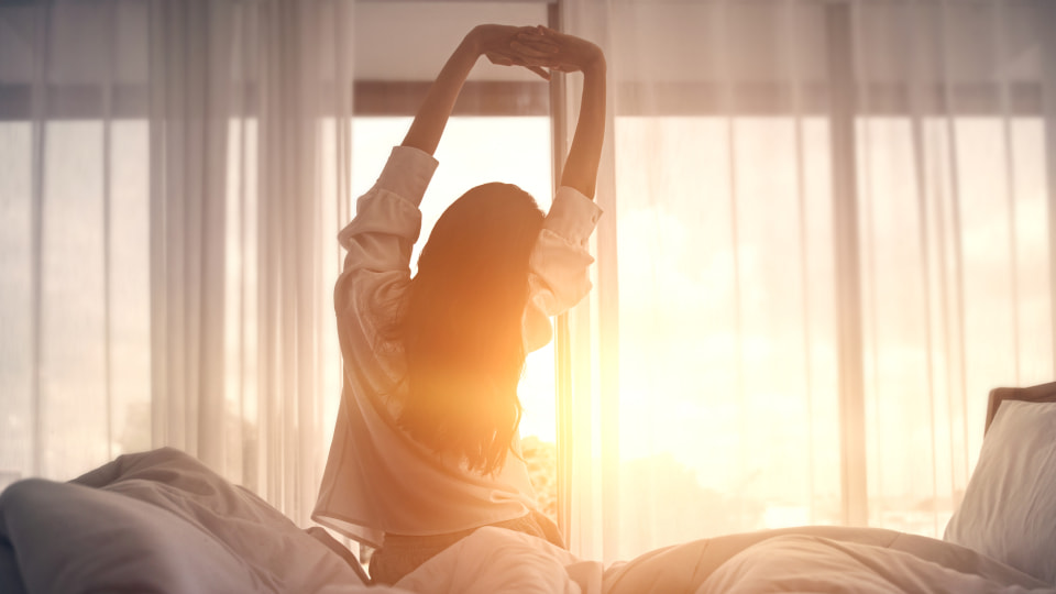 Person sat on the edge of a bed stretching their arms in the air with sun shining through curtains into the room.