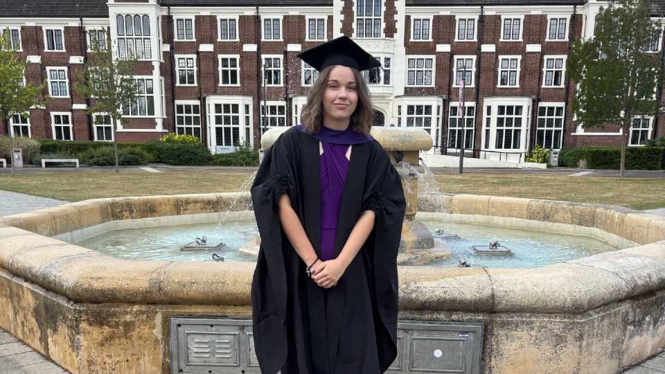 Daisy Newton wearing a graduation cap and gown, standing in front of the Hazlerigg fountain on campus.
