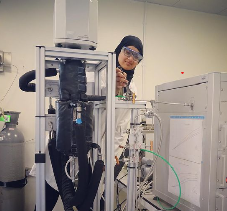 Photo of Humera wearing a lab coat and goggles stood behind a machine in a lab environment