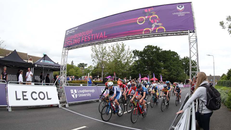 A group of cyclists cycling under a big banner reading 'Loughborough Cycling Festival' with spectators watching..