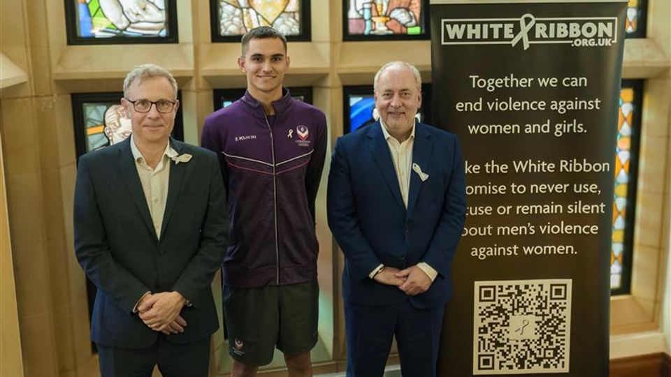 Chief Operating Officer Richard Taylor, Vice-Chancellor Professor Nick Jennings and student Henry Hughes standing in front of stained glassed windows with a White Ribbon standing banner.